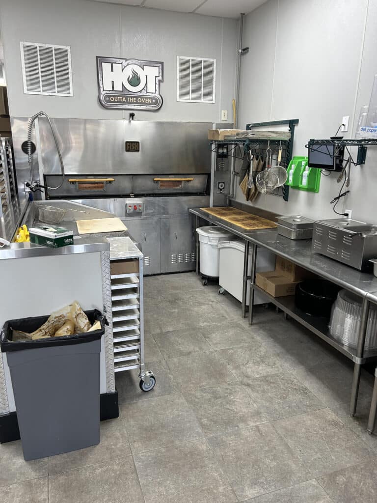 Clean commercial kitchen with stainless steel equipment, prep area, and oven labeled "FISH" under a "HOT OUTTA THE OVEN" sign