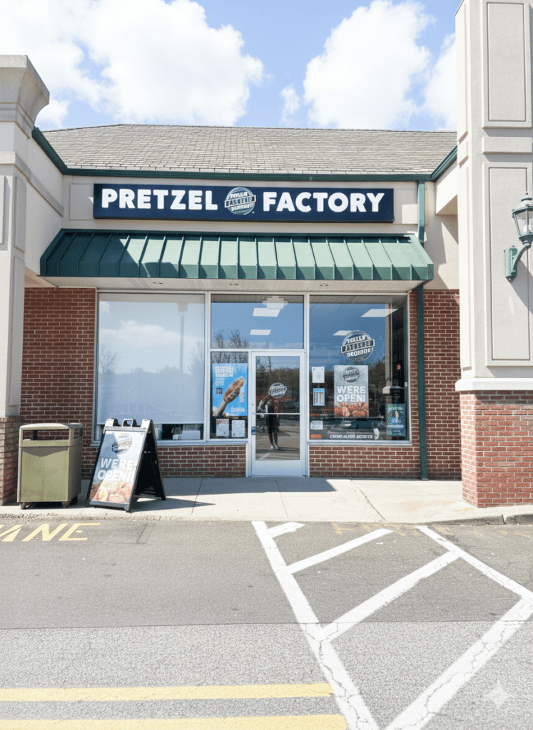 Front view of a Pretzel Factory storefront with a green awning, large windows, and a glass door, displaying “We’re Open” signs and promotional posters, set in a small shopping plaza under a bright, partly cloudy sky.
