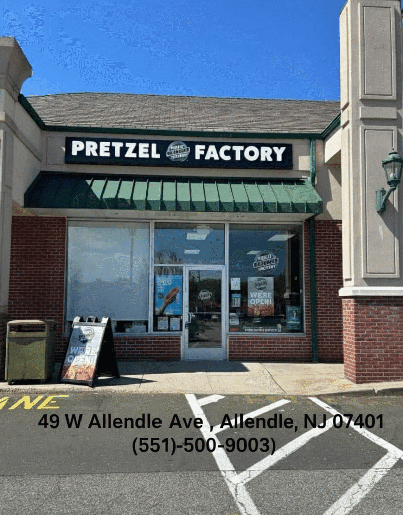 Exterior of a Pretzel Factory storefront with a green awning and large sign reading “Pretzel Factory,” glass entrance doors, and sidewalk signage in a small shopping plaza on a sunny day.